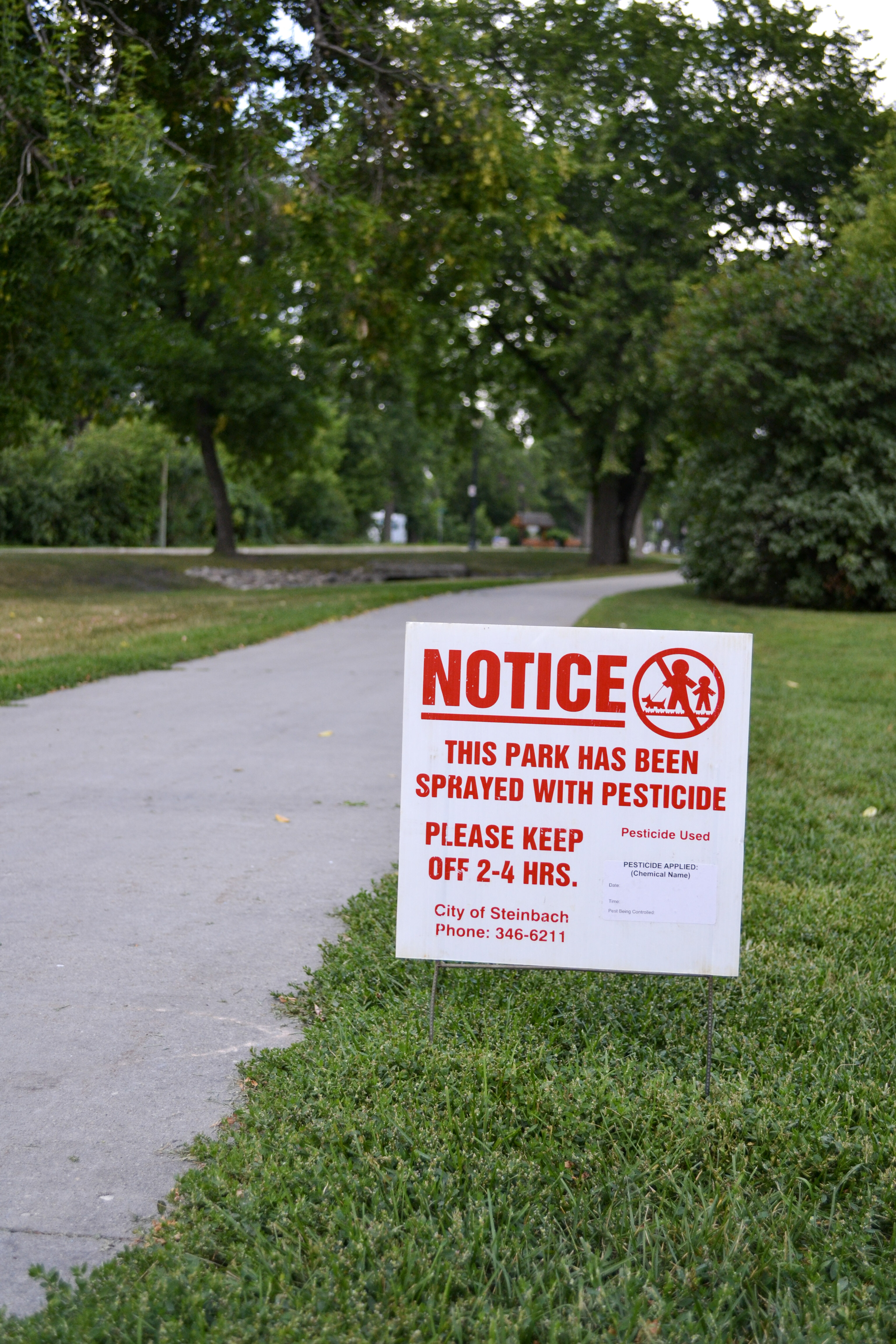 City of Steinbach Notice of Pesticide in-ground sign that reads, in bright red text, "Notice \ This park has been sprayed with pesticide. \ Please keep off 2-4 hrs. \ City of Steinbach Phone: 346-6211."